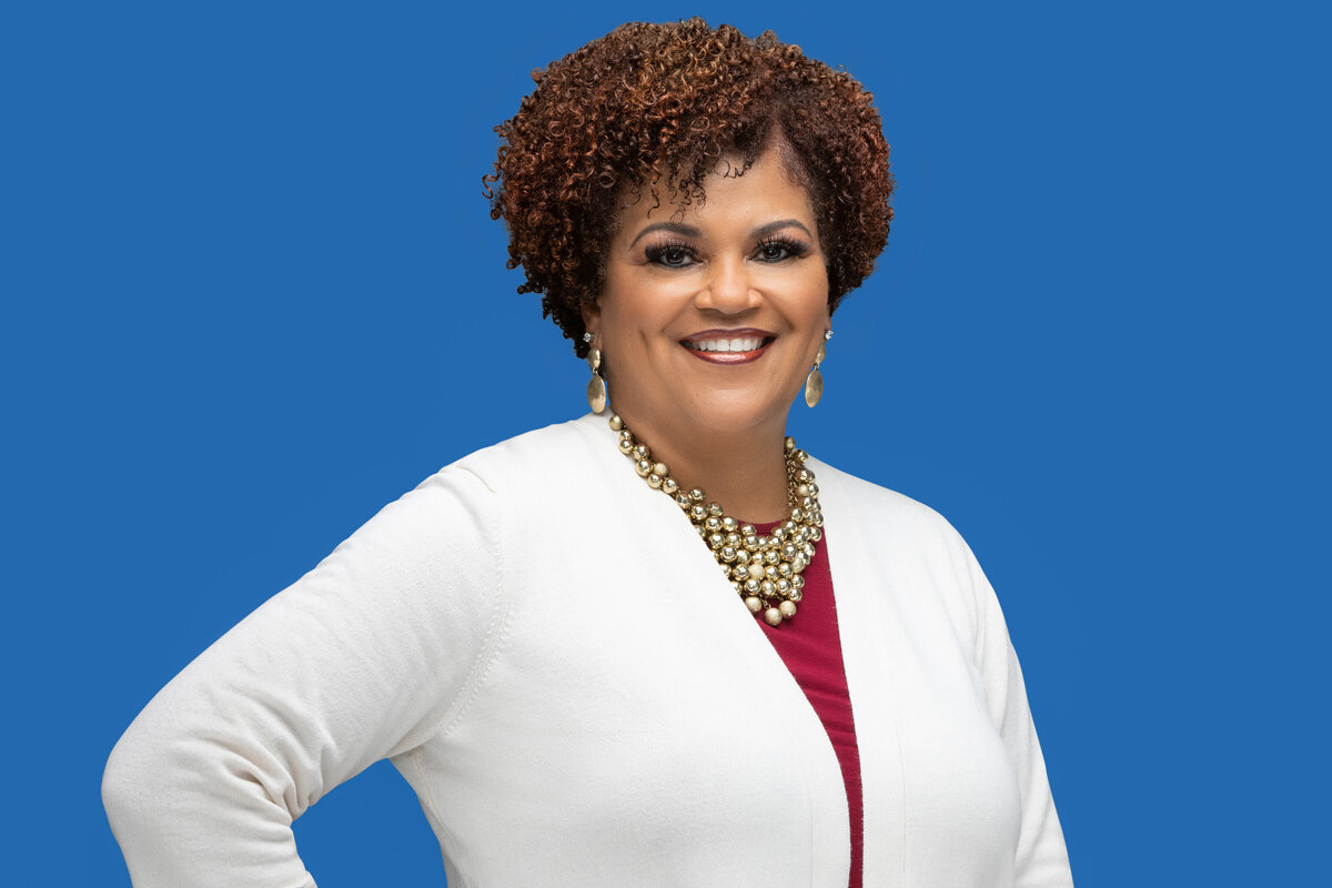 An African American black businesswoman in a white sweater with naturally curly hair poses for a professional headshot photo on a blue backdrop for Janel Lee Photography studios in Cincinnati Ohio