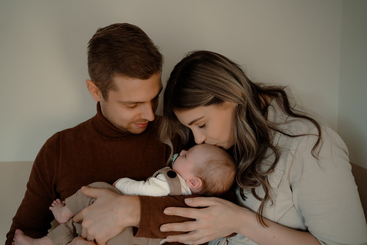 Vader heeft pasgeboren baby op zijn arm en moeder geeft de baby een kusje op zijn voorhoofd tijdens een newbornshoot