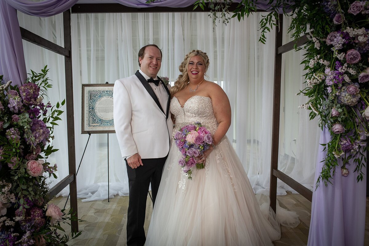 Jewish groom wearing a white tuxedo with a black shawl collar stands beside the Jewish bride with a long blonde braid wearing  a strapless lace wedding gown , holding a cascade bouquet of purple roses, pink roses and purple flowers. The bride and groom are standing under the chuppah which is draped in sheer purple fabric and has two large sprays of purple roses, pink roses, lilac roses and greenery. Behind the bride and groom is the blue and white ketubah is on an easel behind them at The Liff Center, Nashville.