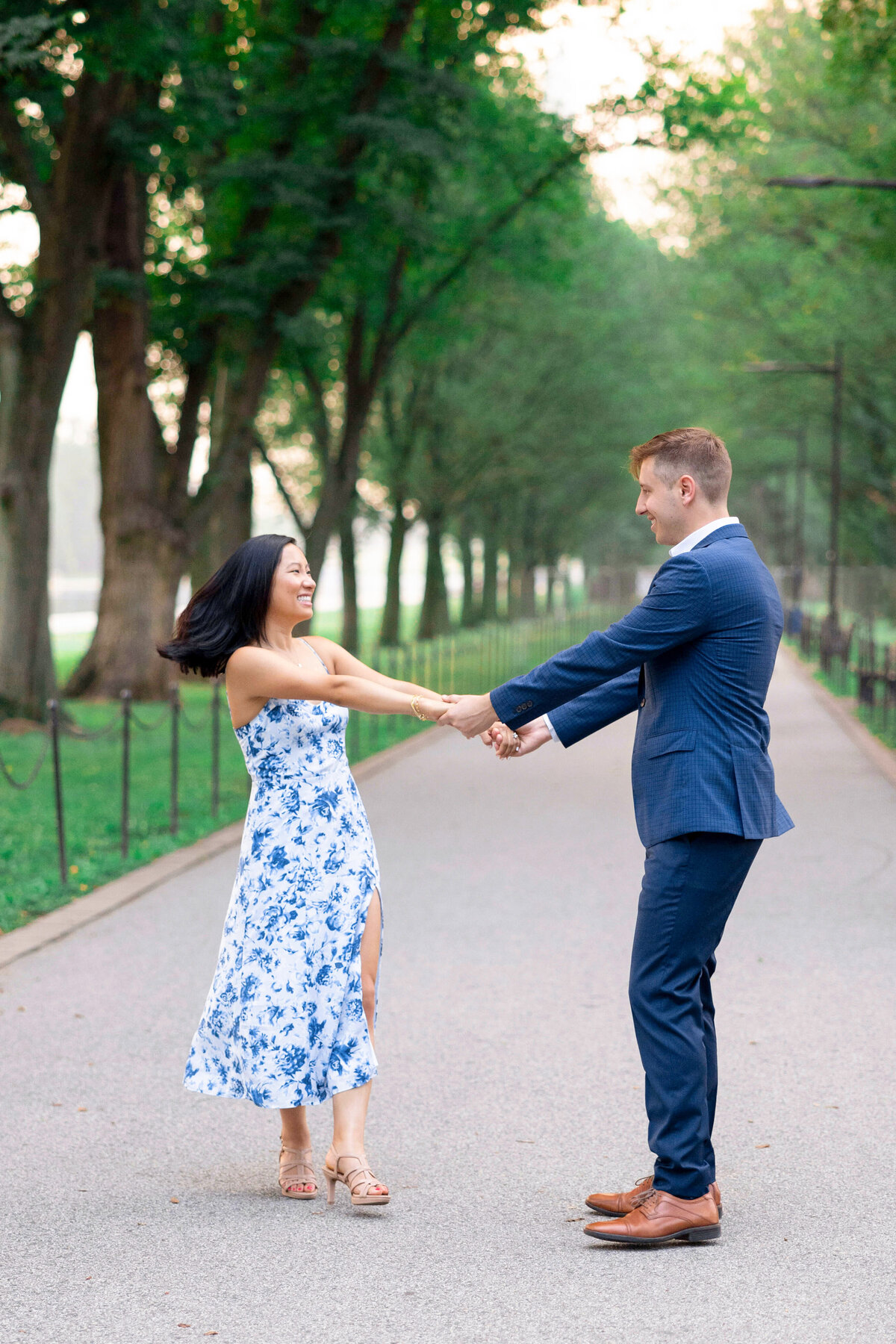 A couple dances joyfully on a tree-lined pathway. The woman is in a blue floral dress, and the man wears a blue suit. They hold hands, surrounded by lush greenery and a serene atmosphere.