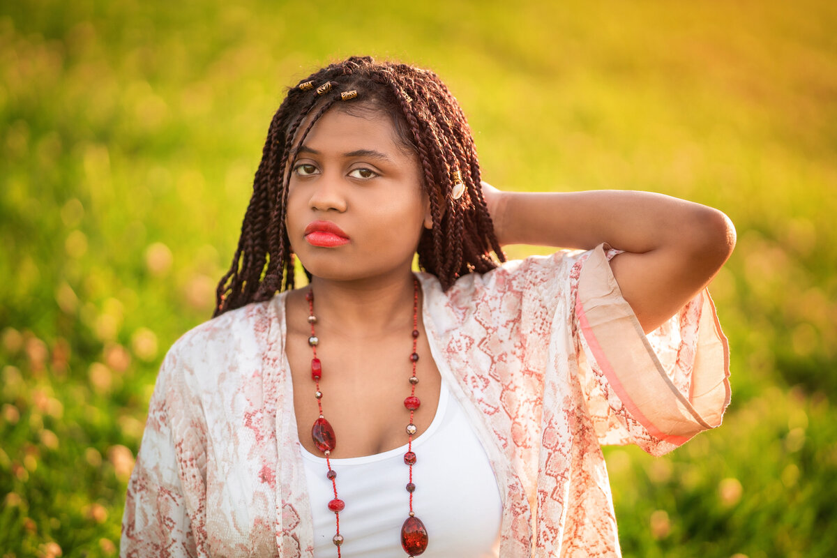 African American girl with long braids.  She is wearing a white tank top and red necklace.  She has a kimono on.  She is looking at the camera with a serious expression and has one hand behind her head.  The background is grassy with lots of warm light showing through.