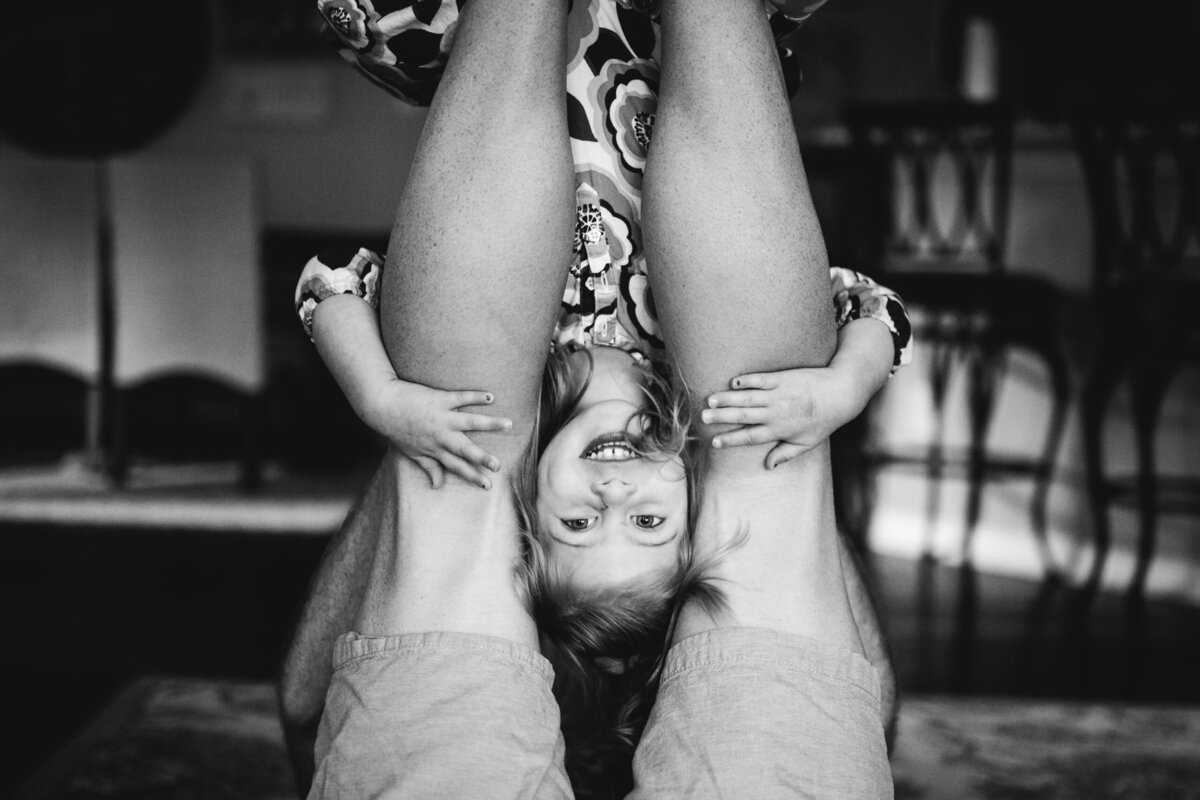 a black and white image of a young girl upsade down on her father's legs.