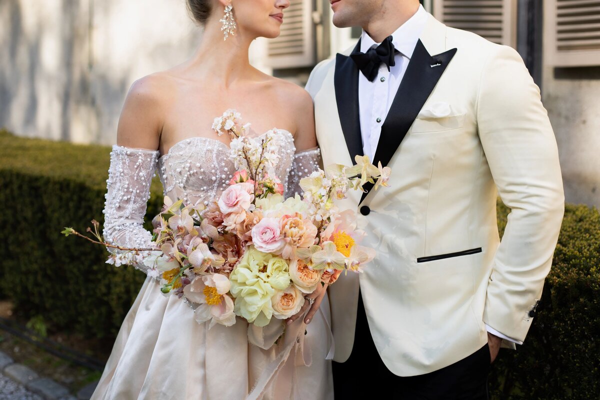 Wedding Photographers in RI | Kelsey Sheehan Photography Timeless Rhode Island Weddings | Bride and groom facing each other. Bride in an off-shoulder gown holding a colorful bouquet. Groom in a white tuxedo with black accents, conveying elegance and romance.