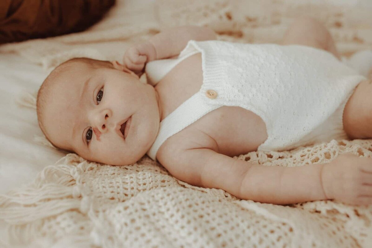 A baby wearing a white knitted romper lies on a beige textured blanket, looking up with one arm bent near the head.