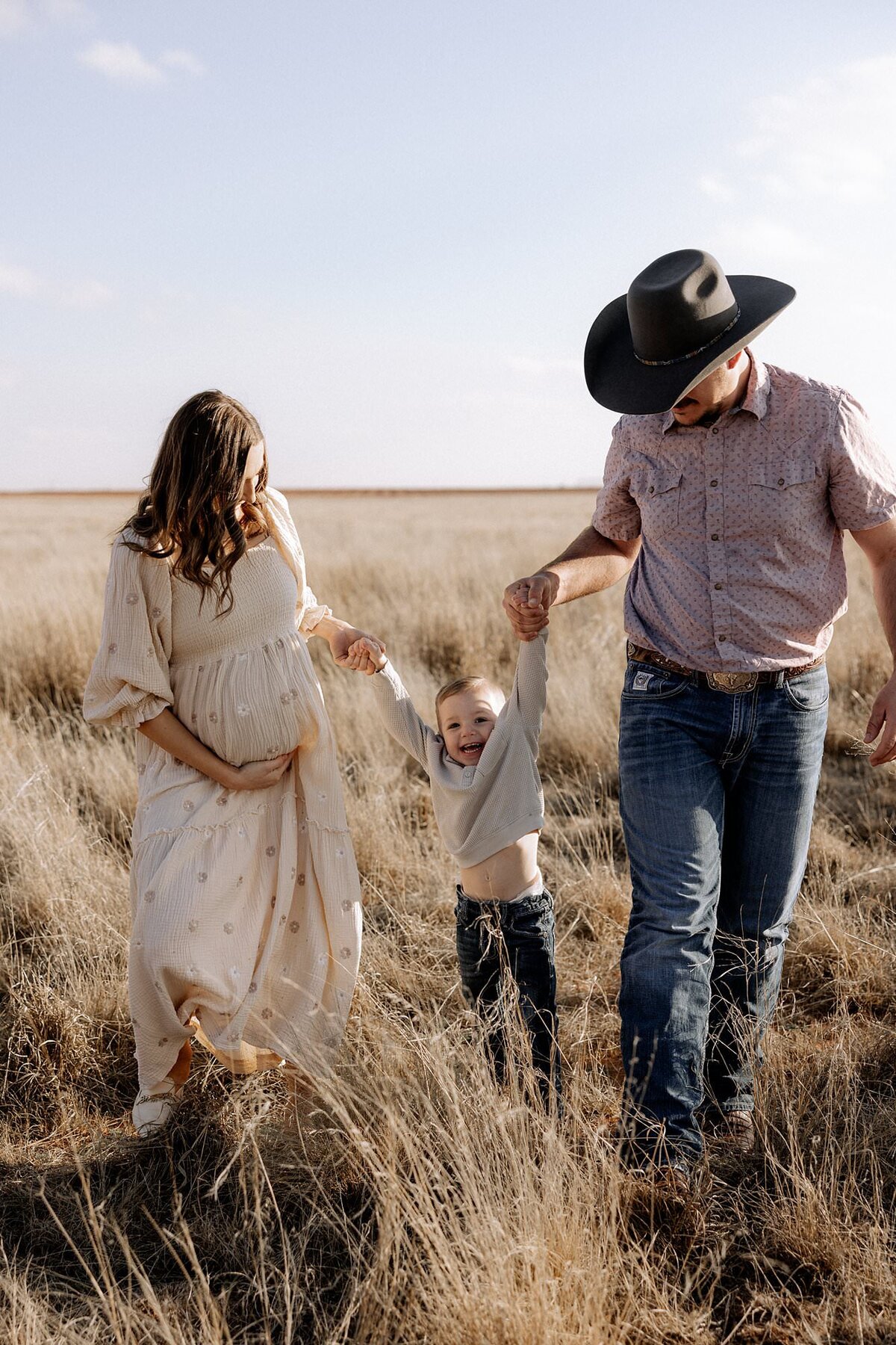 maternity photo of mom and dad swinging two year old son by his arms in an open grass field