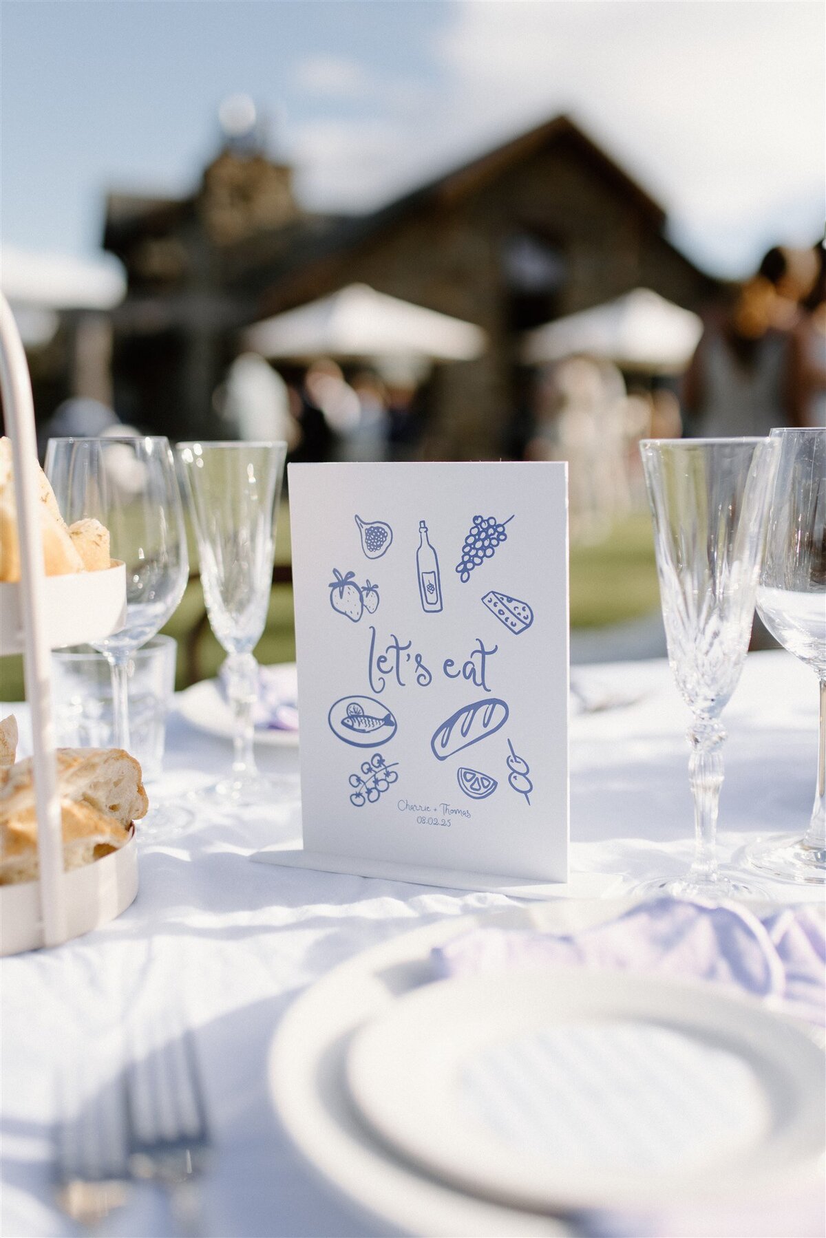 This image captures the same charming “Let’s Eat” menu card but from another table, with the historic stone house blurred behind it. You can almost hear the laughter and clinking glasses that follow an outdoor feast.