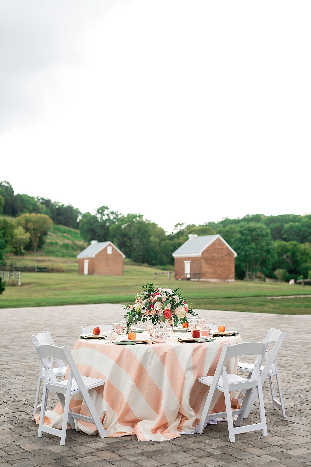 A reception table with a peach and white striped table cloth and white garden chairs in the courtyard at Ravenswood mansion is topped with a large floral centerpiece in a gold footed bowl with greenery, peach, blush and pink flowers and mismatched vintage china with fresh peaches at each place setting.
