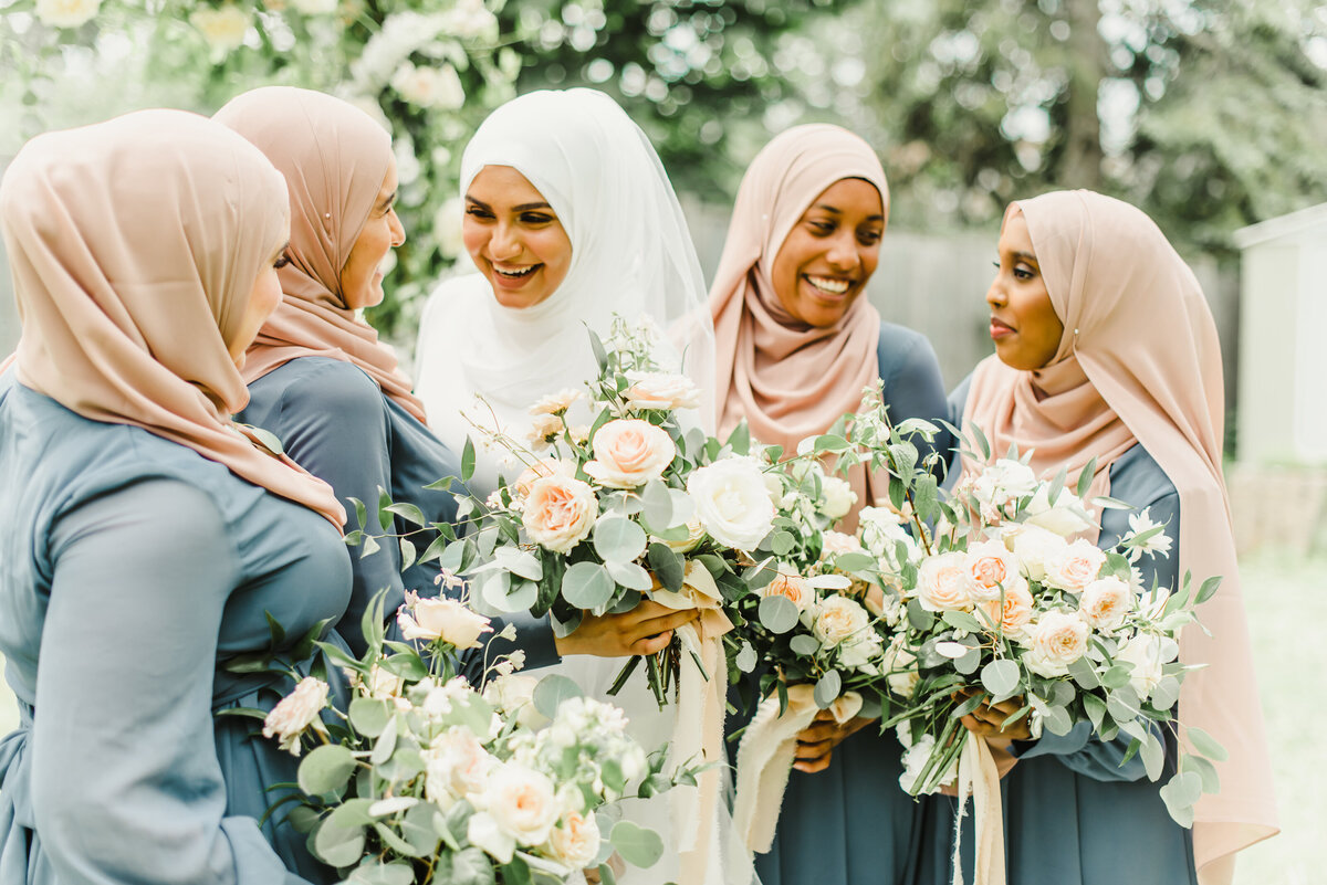 A Muslim bride and her four bridesmaids in blush hijabs and dusty blue dresses smile at each other while showing off their large bouquets filled with white, cream and blush garden roses.
