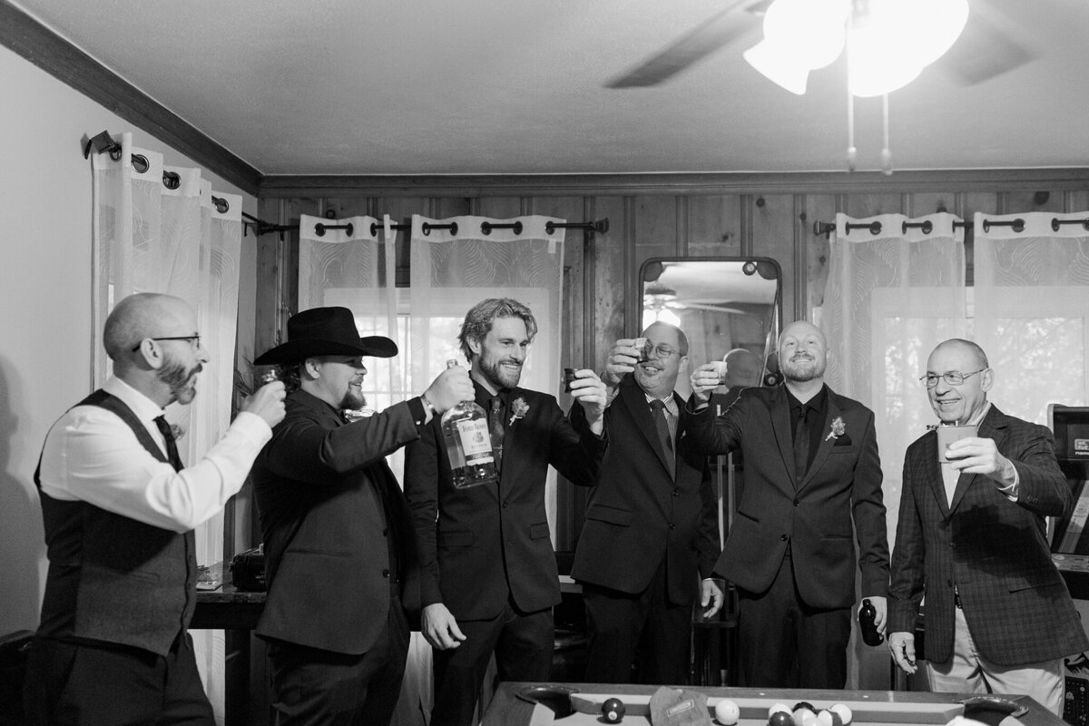 A black and white candid shot of a groom and his groomsmen getting ready to take a shot together before a wedding in Dallas, Texas. They are all wearing suits and holding up their shot glasses in celebration.