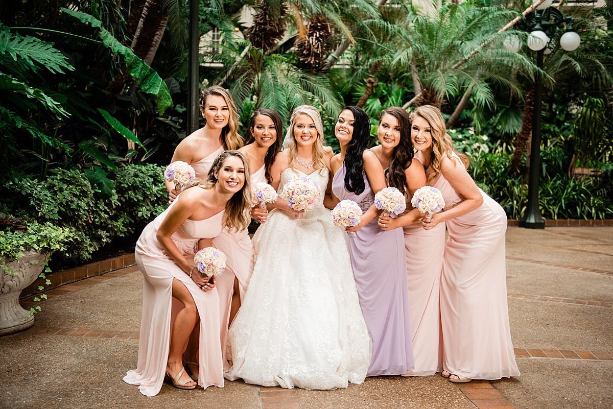Bridesmaids surrounding her bride and smiling at camera, they are wearing soft pink and purple shades