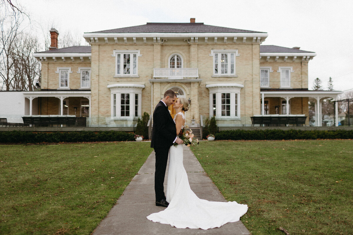 A bride and groom stand facing each other on a pathway at Cape Picton, holding hands, in front of a large, historic building with a lawn, as captured by an exceptional wedding photographer in Toronto.
