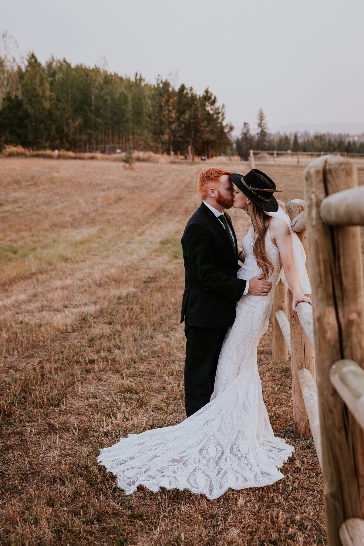 Bride and groom kiss while leaning against farm fence.