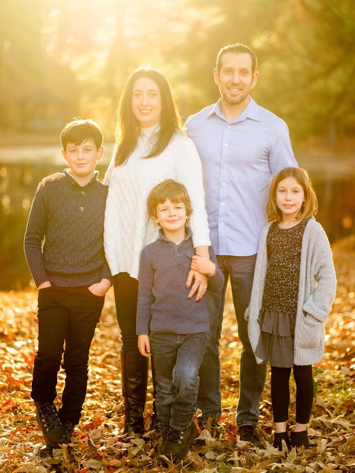Parents with three kids standing on a wooded area covered in leaves.