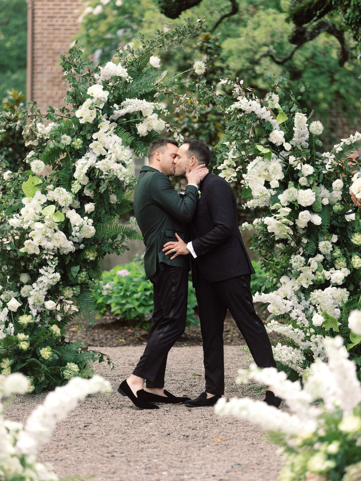 In a romantic outdoor setting, two grooms share a tender kiss, symbolizing love and commitment while surrounded by lush floral arrangements.