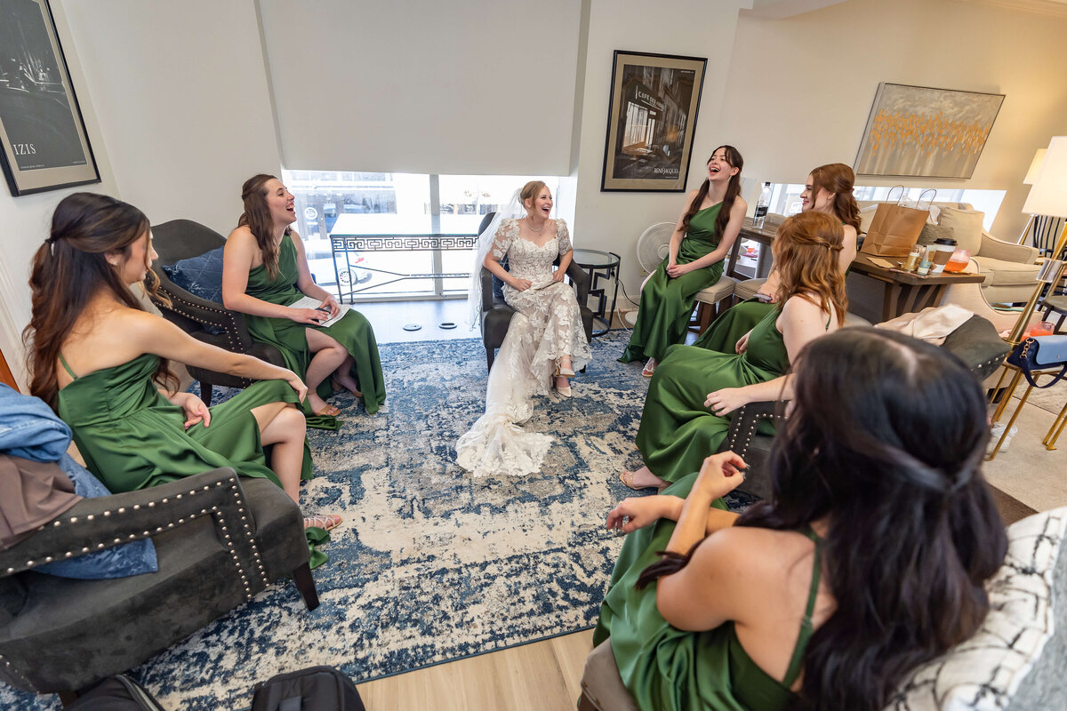 A bride in a white dress sits smiling with six bridesmaids in green dresses around her in a cozy, well-lit room. Everyone appears happy and relaxed, sharing a moment before the wedding.