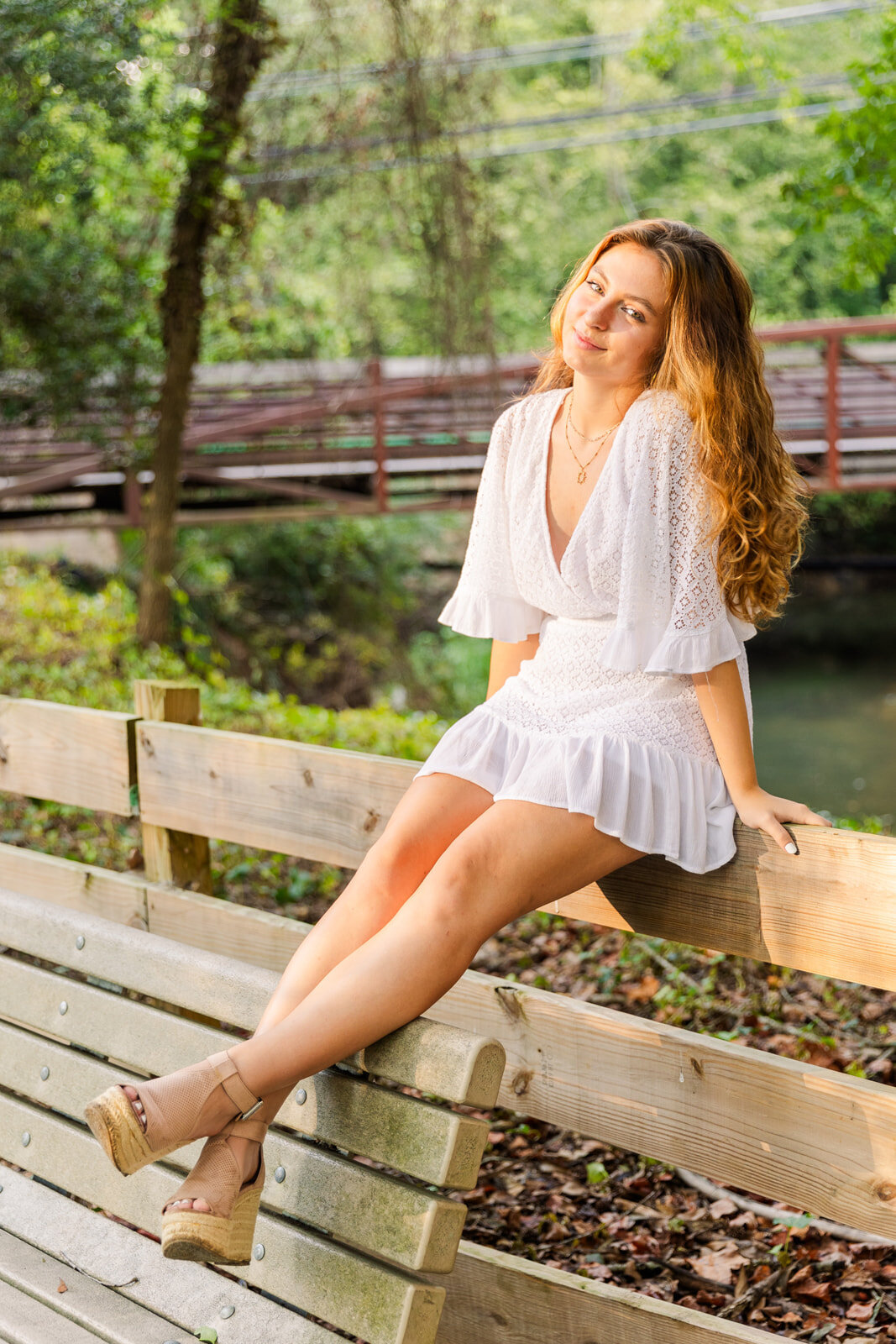 teenage girl wearing a short white dress sitting on a wooden fence in front a a bridge in an Atlanta pack during senior portraits