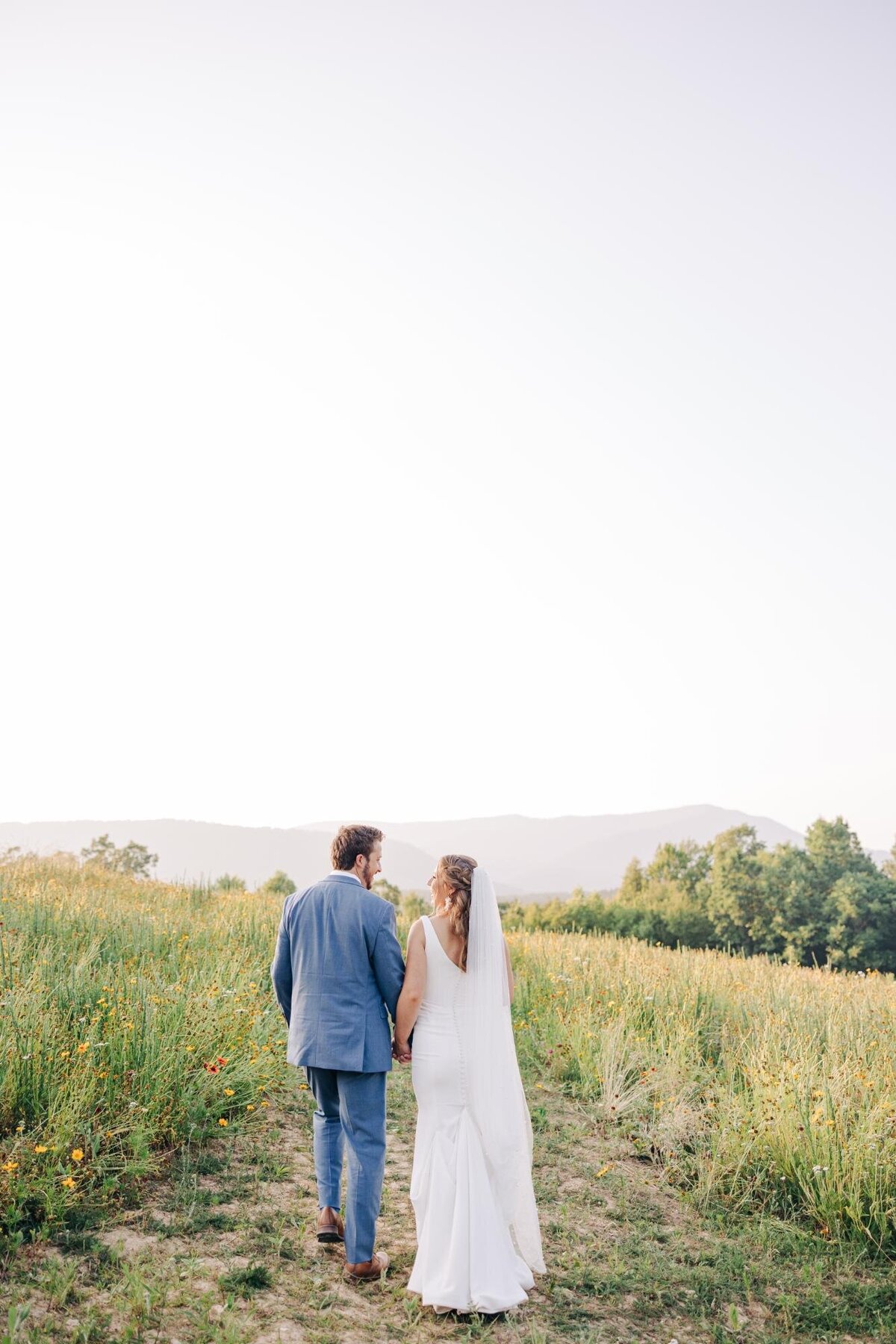 A couple in wedding attire holding hands while walking through a field with a mountainous backdrop.