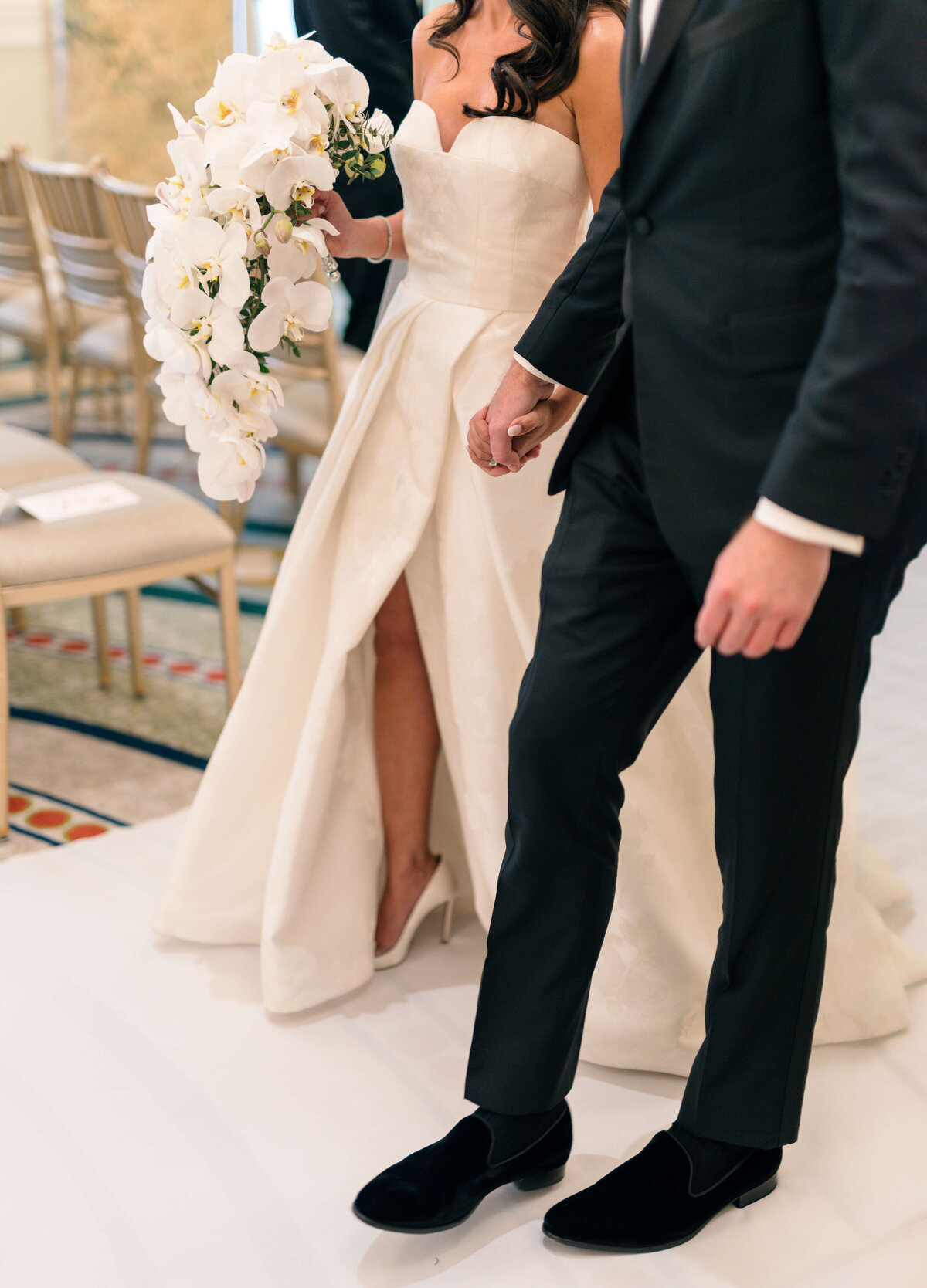 A bride in a white strapless gown holding a bouquet of white flowers walks hand in hand with a groom in a black suit and velvet shoes. They are indoors, surrounded by chairs and a decorative carpet.