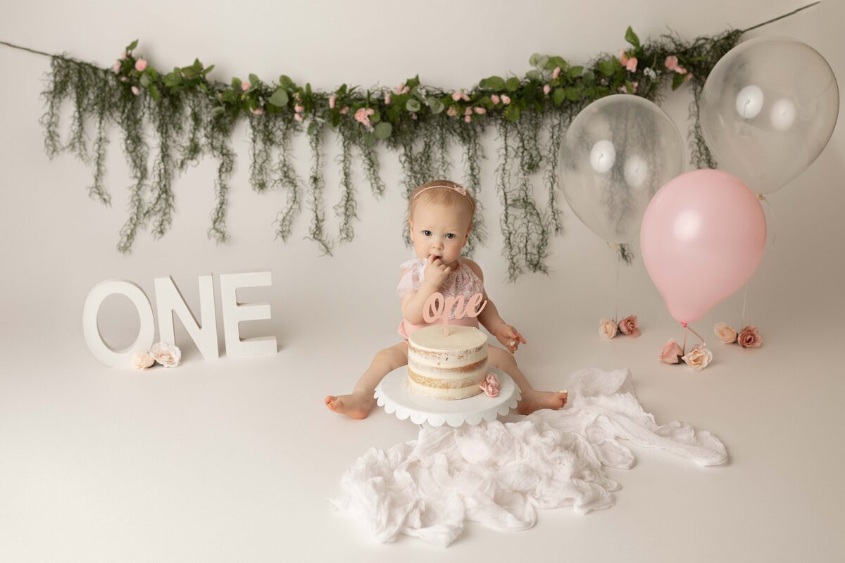 Baby taking their first take of cake during cake smash. Baby is wearing white with white cake adored with a o-n-e cake topper. Greenery draped in the background.