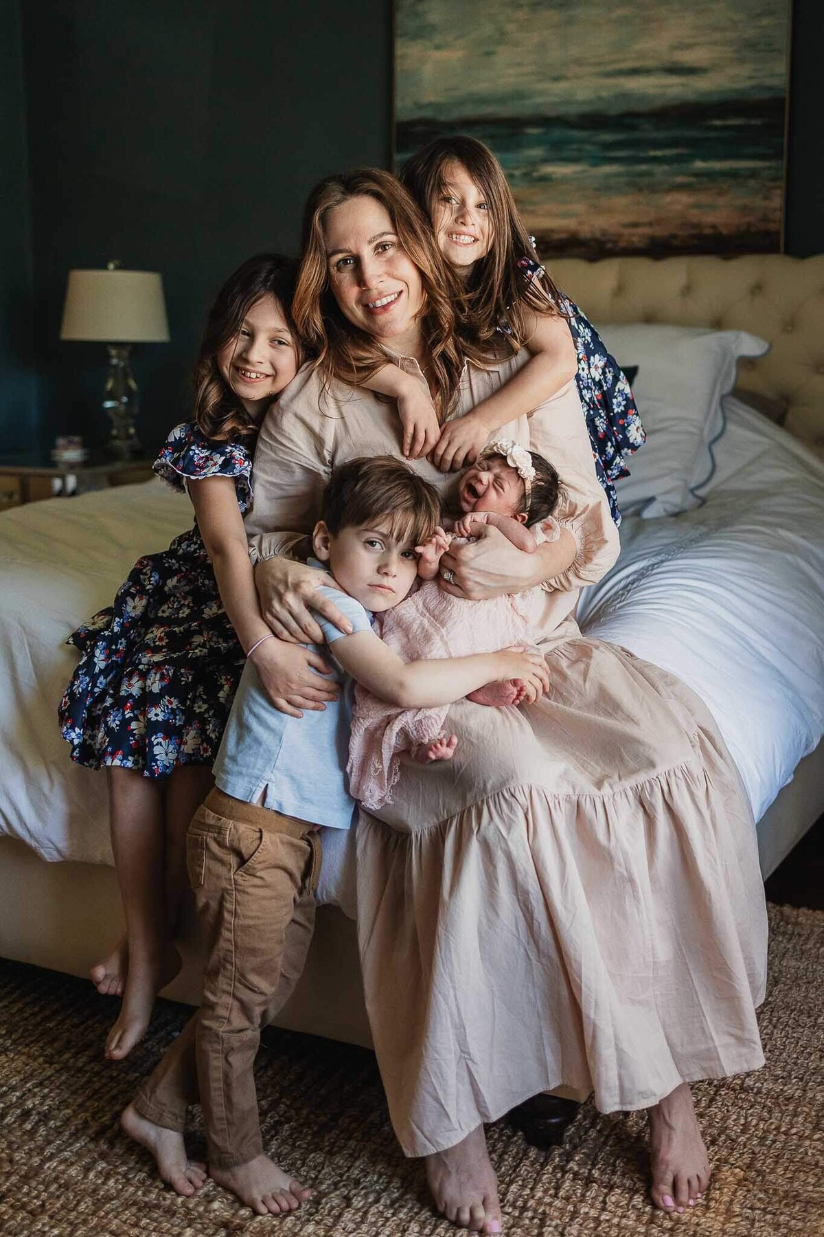 Mom sitting on bed holding newborn daughter while sibling surround her in their Cleveland Heights home.