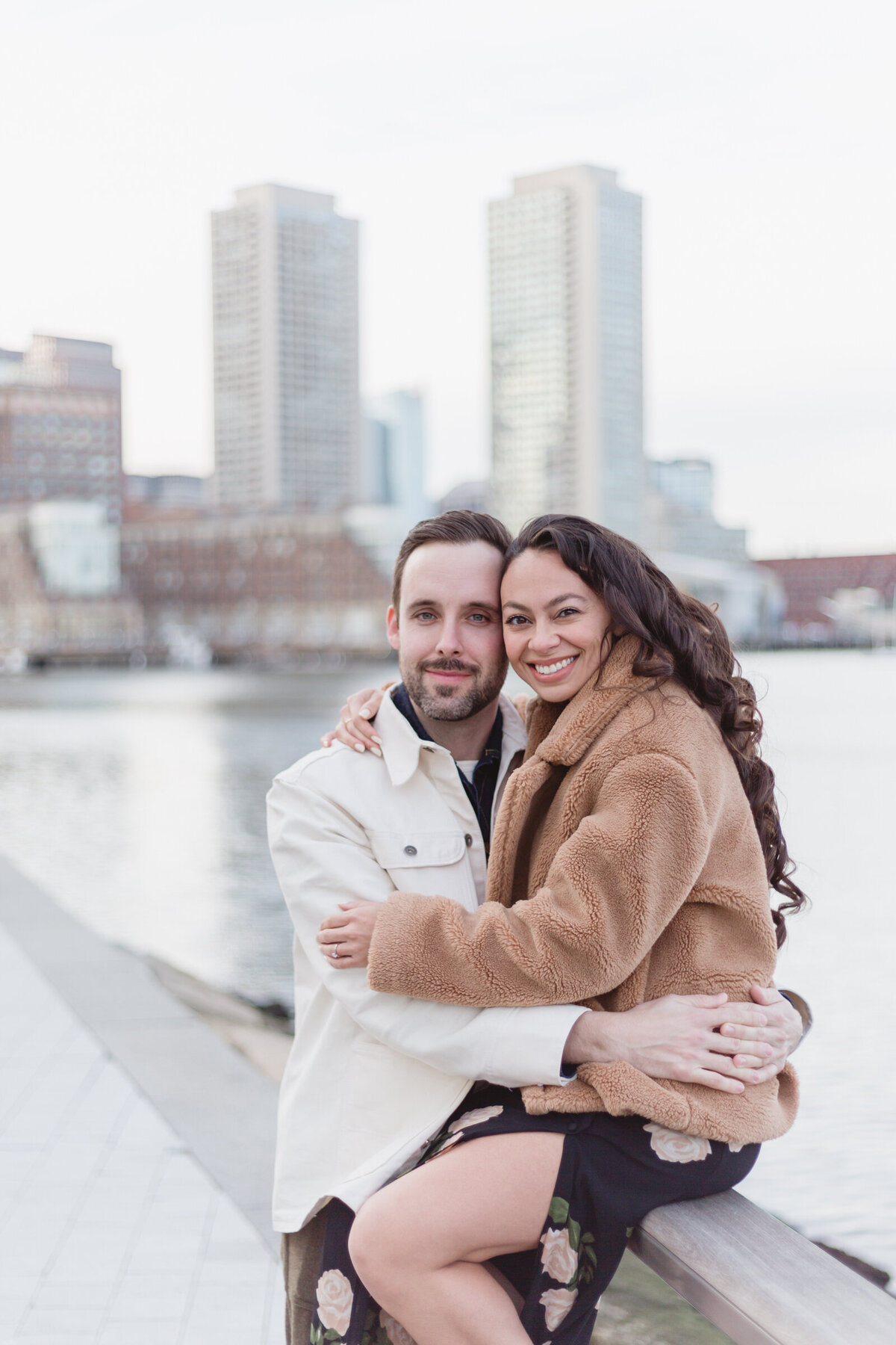 An engaged couple cuddle with the Boston skyline in the background