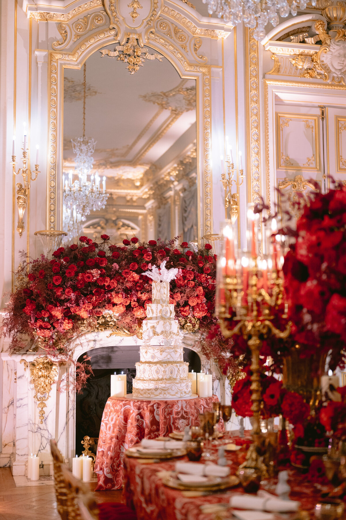 A stunning photo captured by photographer Matteo Coltro at Shangri La Hotel & Ritz Paris Hotel Paris, France, showcases an opulent dining room set for an event. The focal point is a lavish white tiered cake adorned with intricate gold details, adding a touch of extravagance to the room. The overall ambiance exudes luxury with its rich gold and white color scheme, highlighted by grand mirrors, gleaming chandeliers, opulent red floral arrangements, and videography services by Zen Film Works. The table is elegantly set with flickering candles, further enhancing the sophisticated atmosphere.