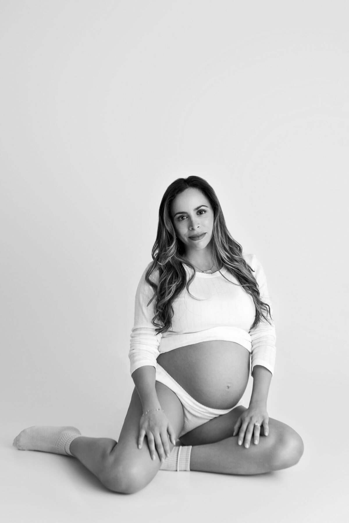 A black-and-white maternity portrait of a pregnant woman sitting on the floor in a relaxed pose. She wears a white long-sleeve top, underwear, and socks, with her bare baby bump visible. Her legs are bent at the knees, and her hands rest gently on her thighs. She gazes directly at the camera with a calm and serene expression, exuding confidence and tranquility. The minimalist background emphasizes the focus on her and the beauty of her pregnancy.