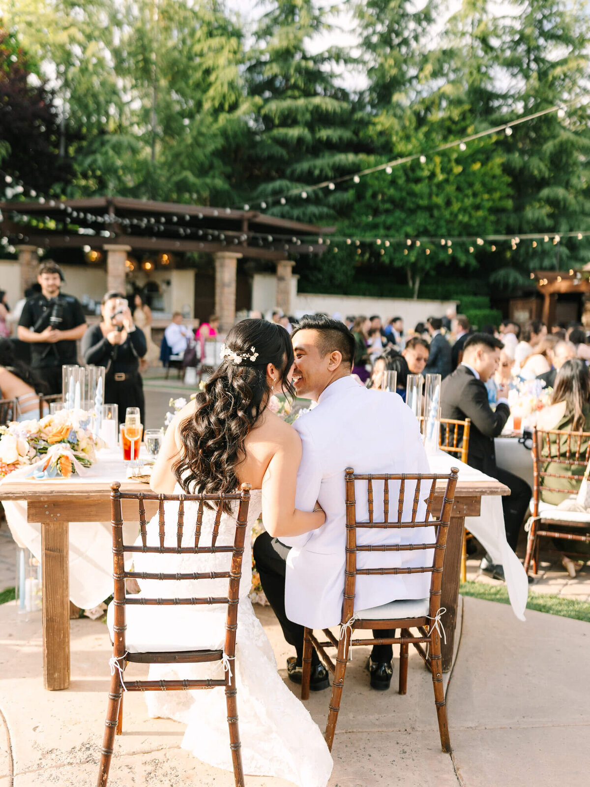 A couple in wedding attire sits closely at a decorated table, surrounded by guests outdoors at Serendipity Garden Weddings Venue. Warm lighting and lush trees create a festive, romantic atmosphere.