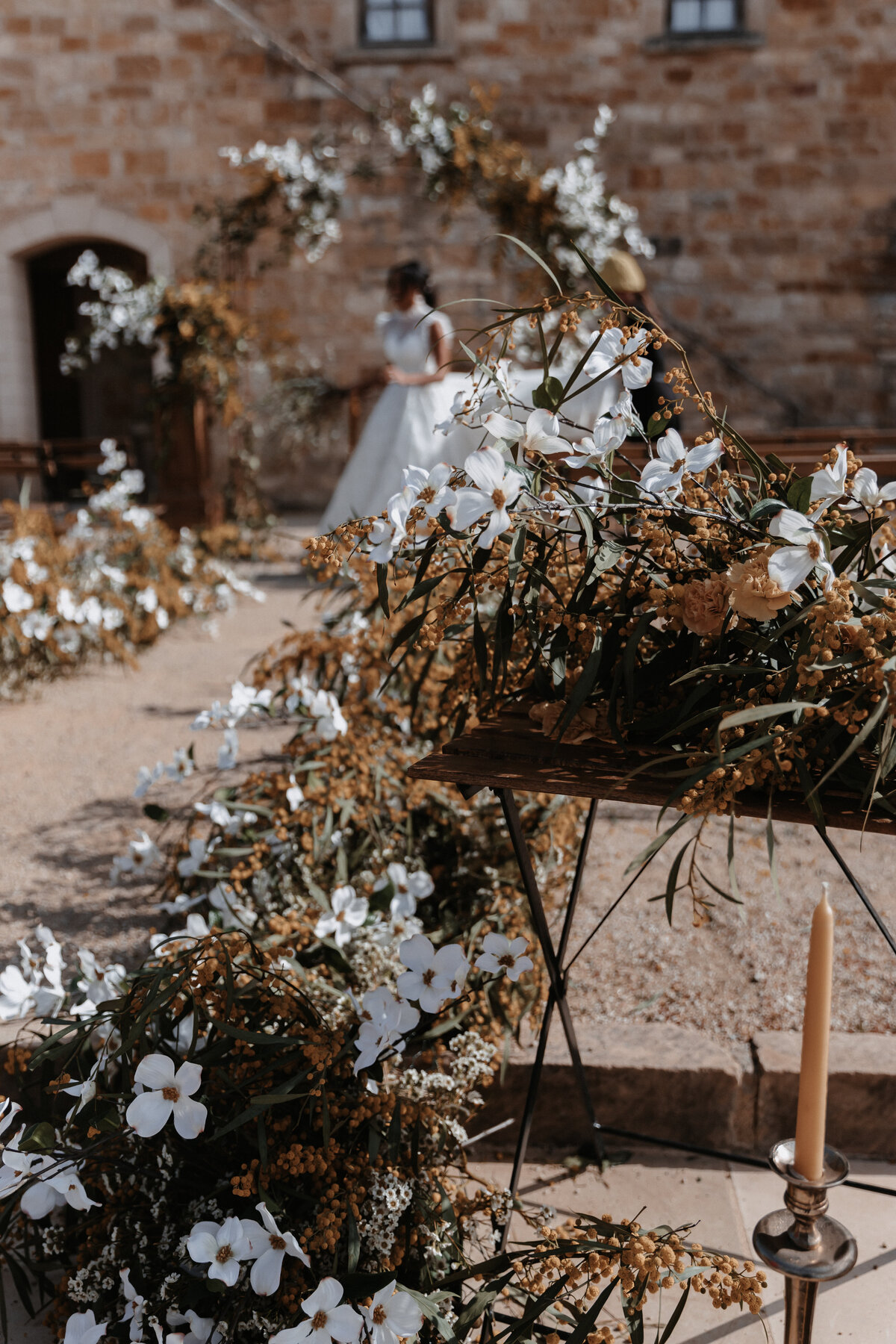 Close-up of lush floral aisle decorations with white flowers and greenery, with the bride visible in the background under a floral arch at Sunstone Winery.