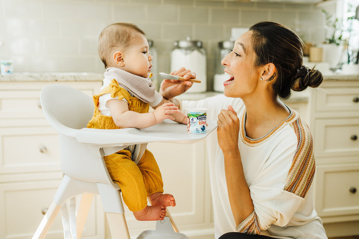 mom feeds baby in highchair yogurt during kids brand photoshoot