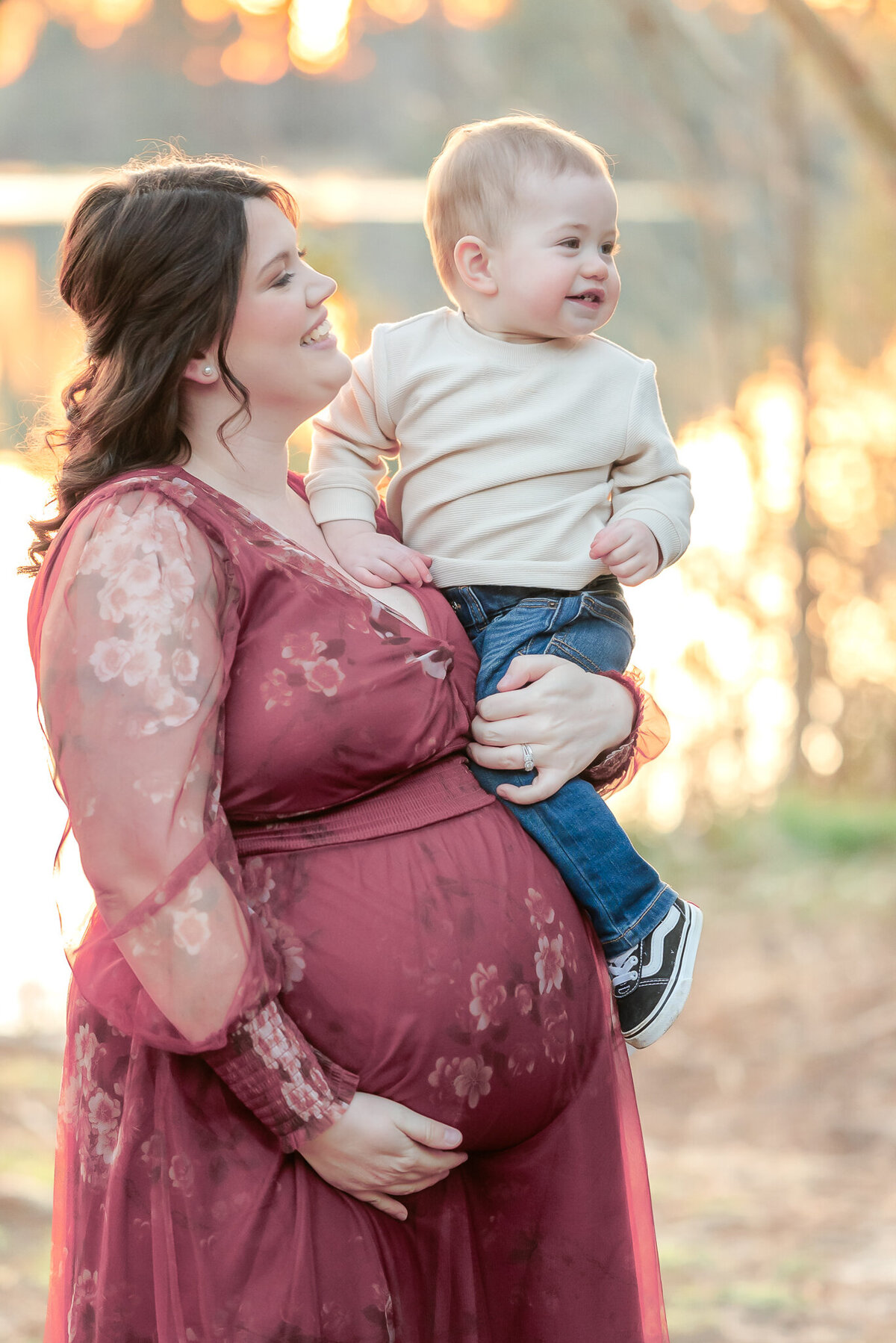 A pregnant woman, wearing a pink floral dress, holds her toddler son in her arms. They are participating in a maternity photography session in Chesapeake, VA.