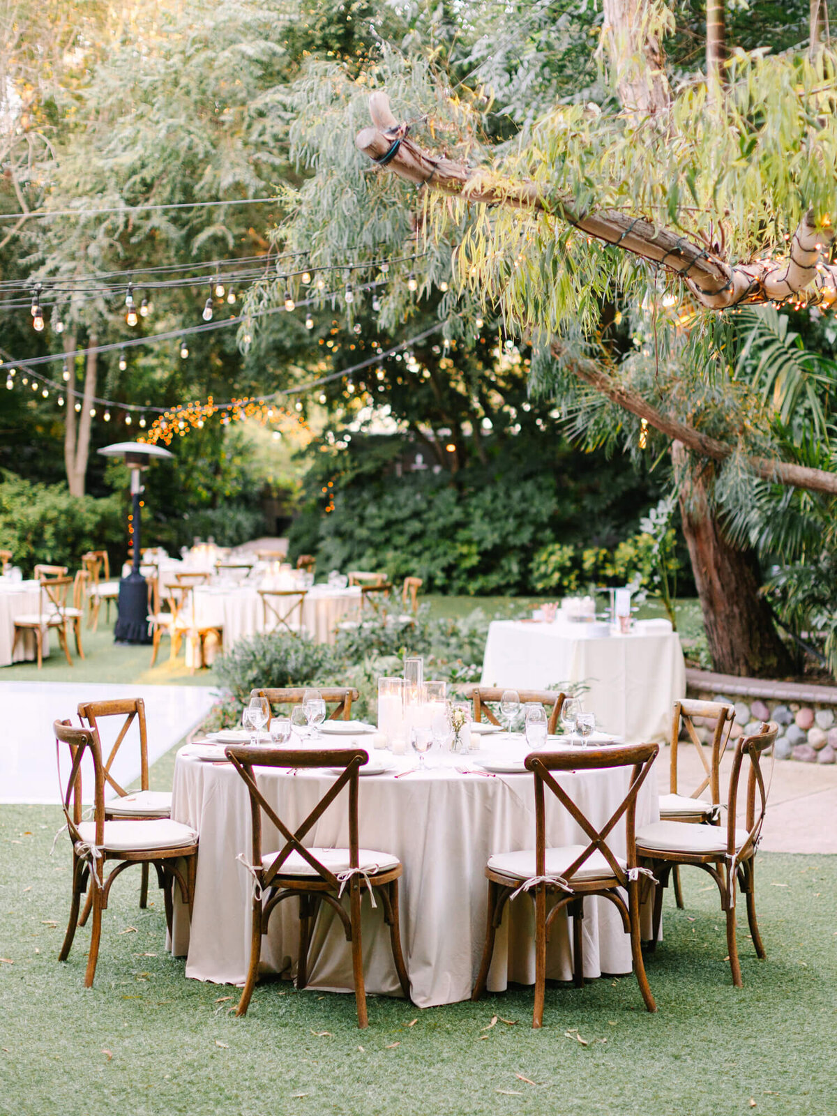 An elegantly set outdoor dining area with wooden chairs, draped tables, and twinkling lights amid lush greenery.