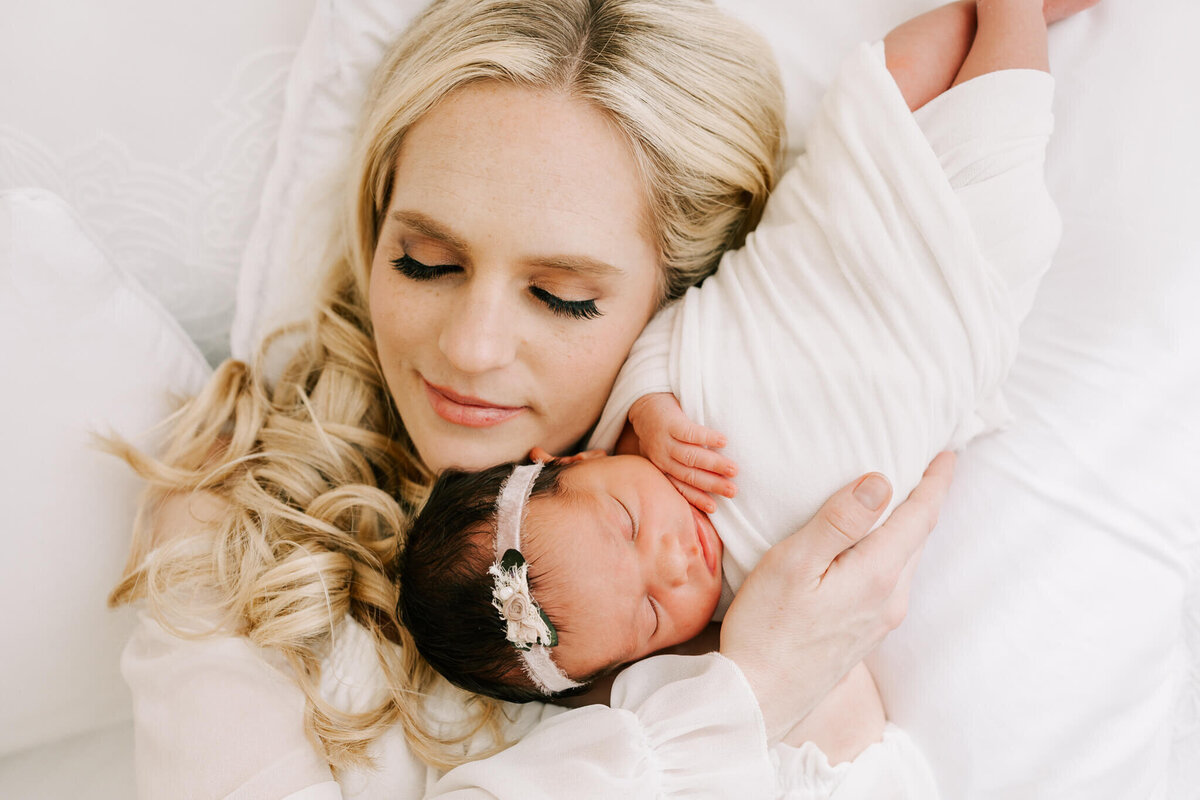 blonde mom holding daughter with her eyes closed. her daughter is wrapped in white wrap
