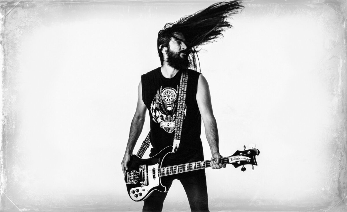 Male musician photo black white Michael Dwyer wearing black t shirt black jeans holding bass guitar while swinging long hair around against white backdrop