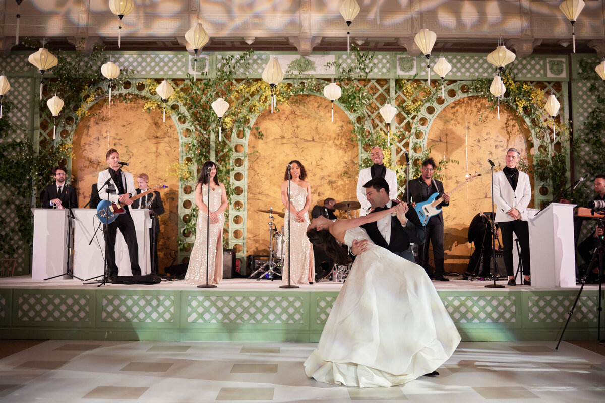 This is a lovely image captured at The New York Public Library in New York City by photographer Shawn Connell. In the photo, a couple is dancing in front of a live band during a joyful celebration. The bride looks radiant in a beautiful white gown, while the groom looks dashing in his formal attire as he gracefully dips her. The band, also dressed elegantly, performs on a decorated stage adorned with greenery and twinkling hanging lights, adding to the magical atmosphere of the event. Additionally, Aaron Navak Films provided videography services, capturing the special moments of the occasion.
