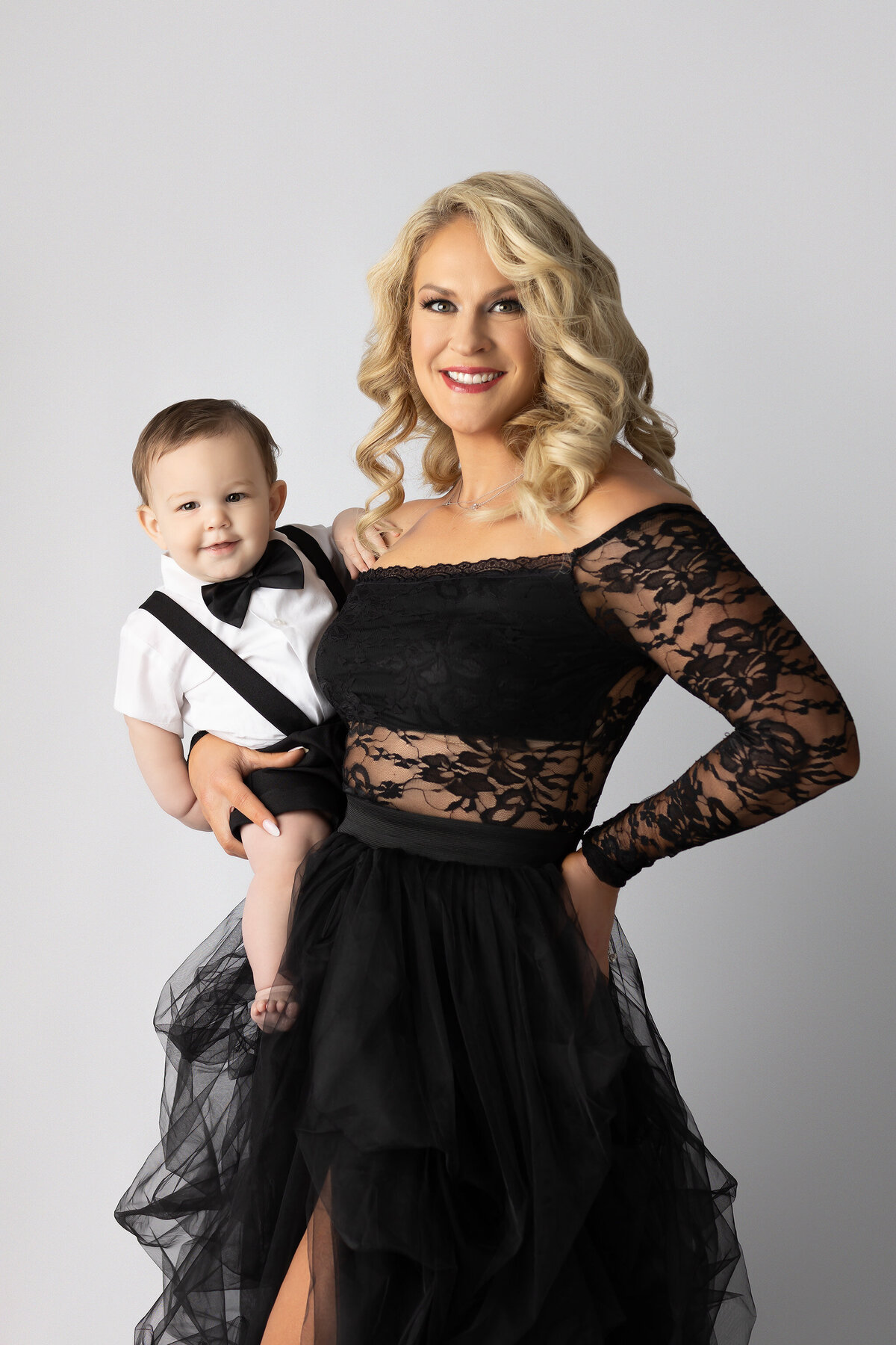 Dallas milestone photographer captures a radiant mother in a sheer black lace gown holding her baby boy dressed in classic black suspenders and bow tie.