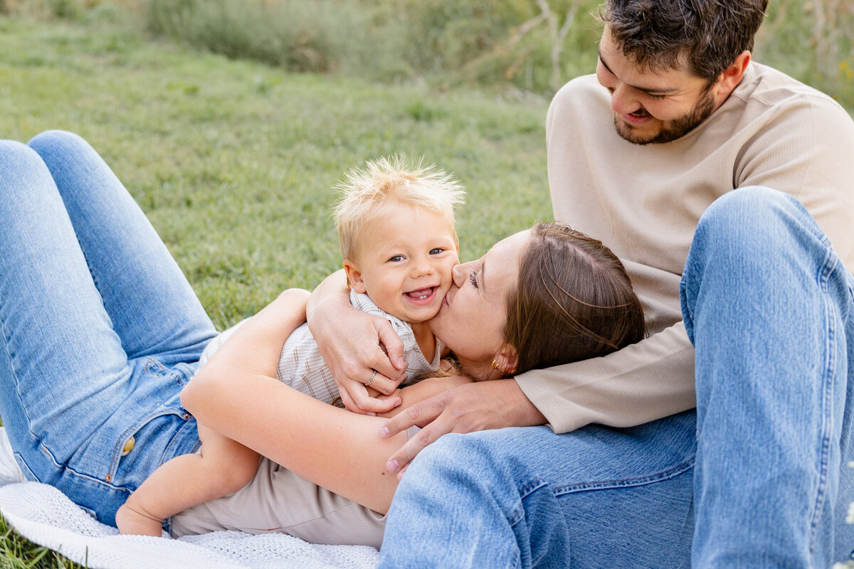  A mother embraces her baby while lying in the grass, with the father lovingly watching over them. This precious family moment was captured by Cassie Lee Photography in Eagle Mountain, Utah.