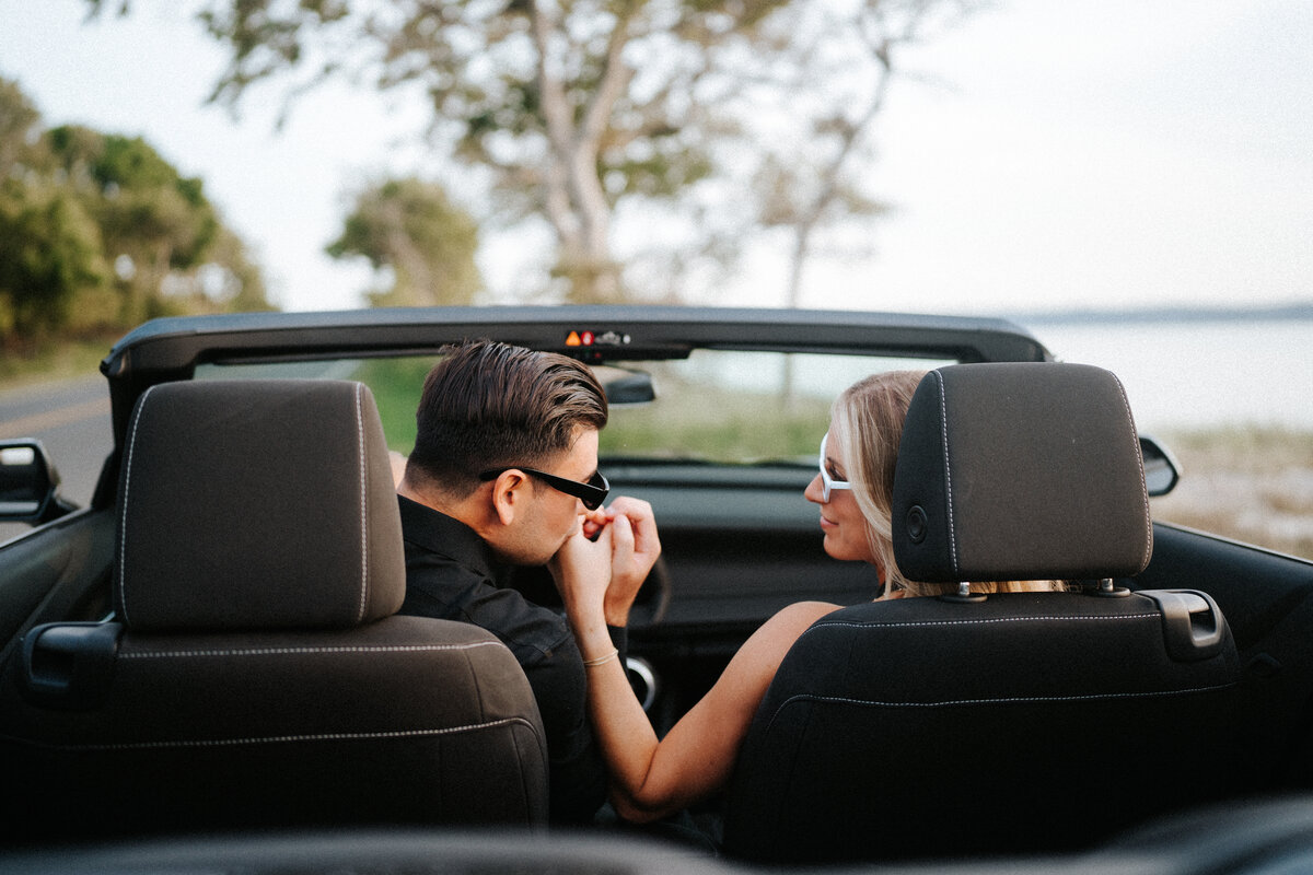 Couple sitting in a convertible at Sandy Hook Beach in New Jersey. The guy is kissing his fiance's hand as the photo is captured from behind the car