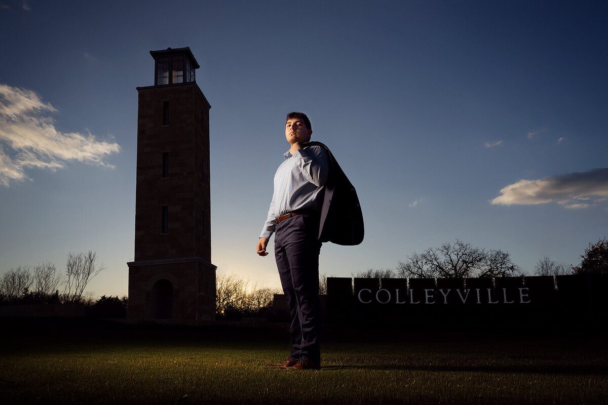 A senior stands confidently with a suit jacket over his shoulder in front of Colleyville Tower at sunset, with the Colleyville sign in the background.