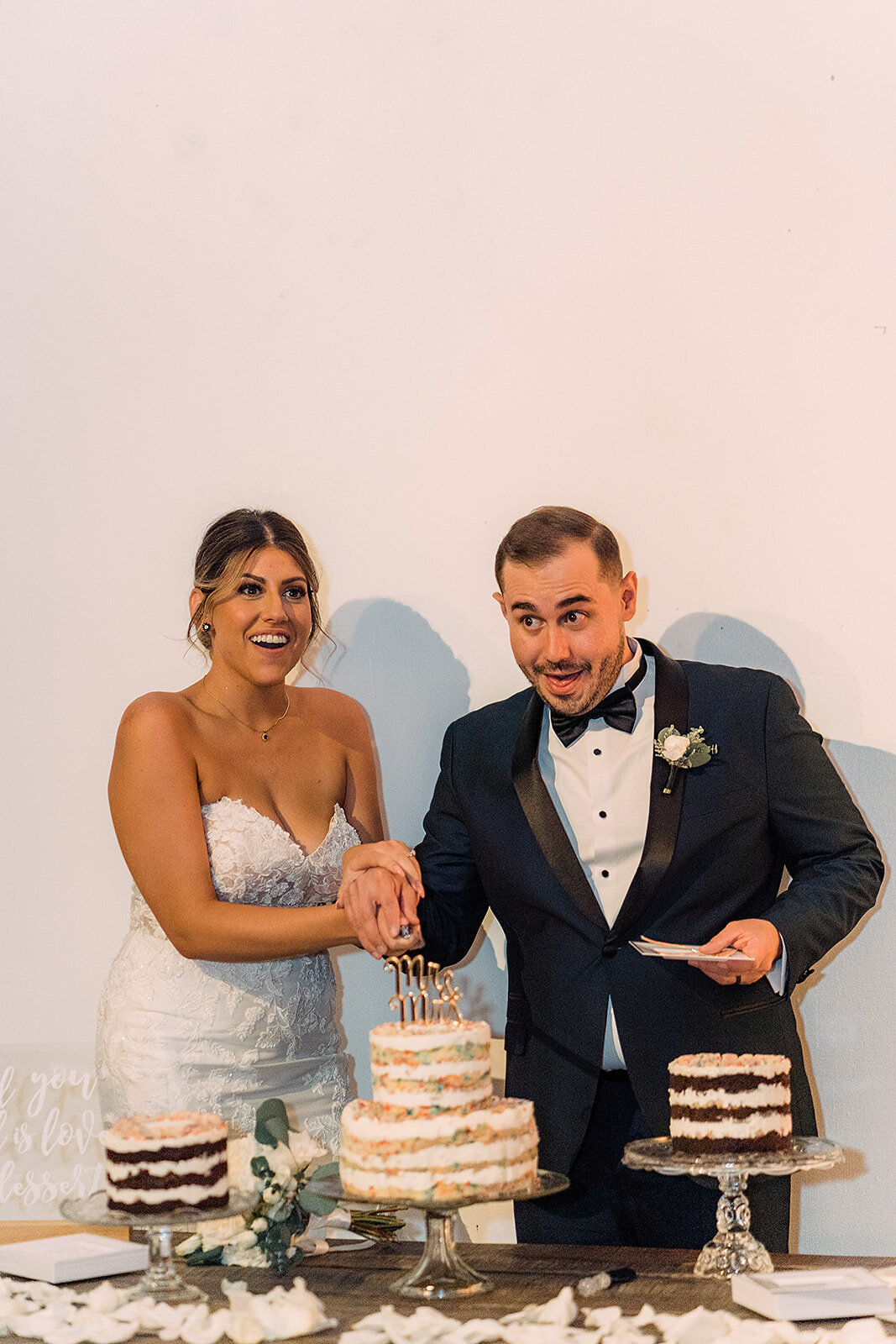 The bride and groom smile as they cut a multi-tiered, naked-style wedding cake with a “Mr & Mrs” topper, surrounded by additional cakes and a display of flowers.