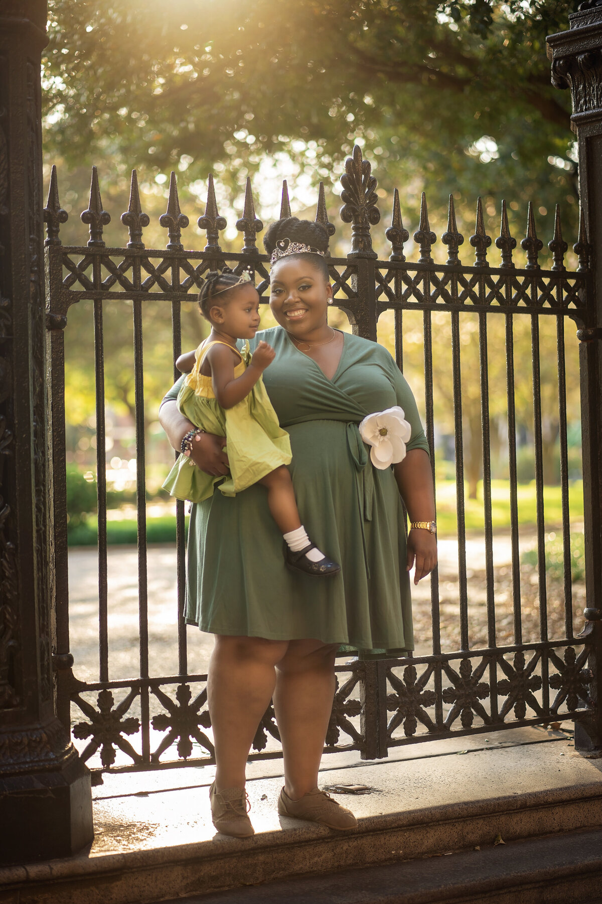 Mommy and daughter dressed like Princess and the Frog standing in front of the gates of Jackson Square.  They are both wearing green and mom has a magnolia on her waist.  The sun is behind them and glowing.