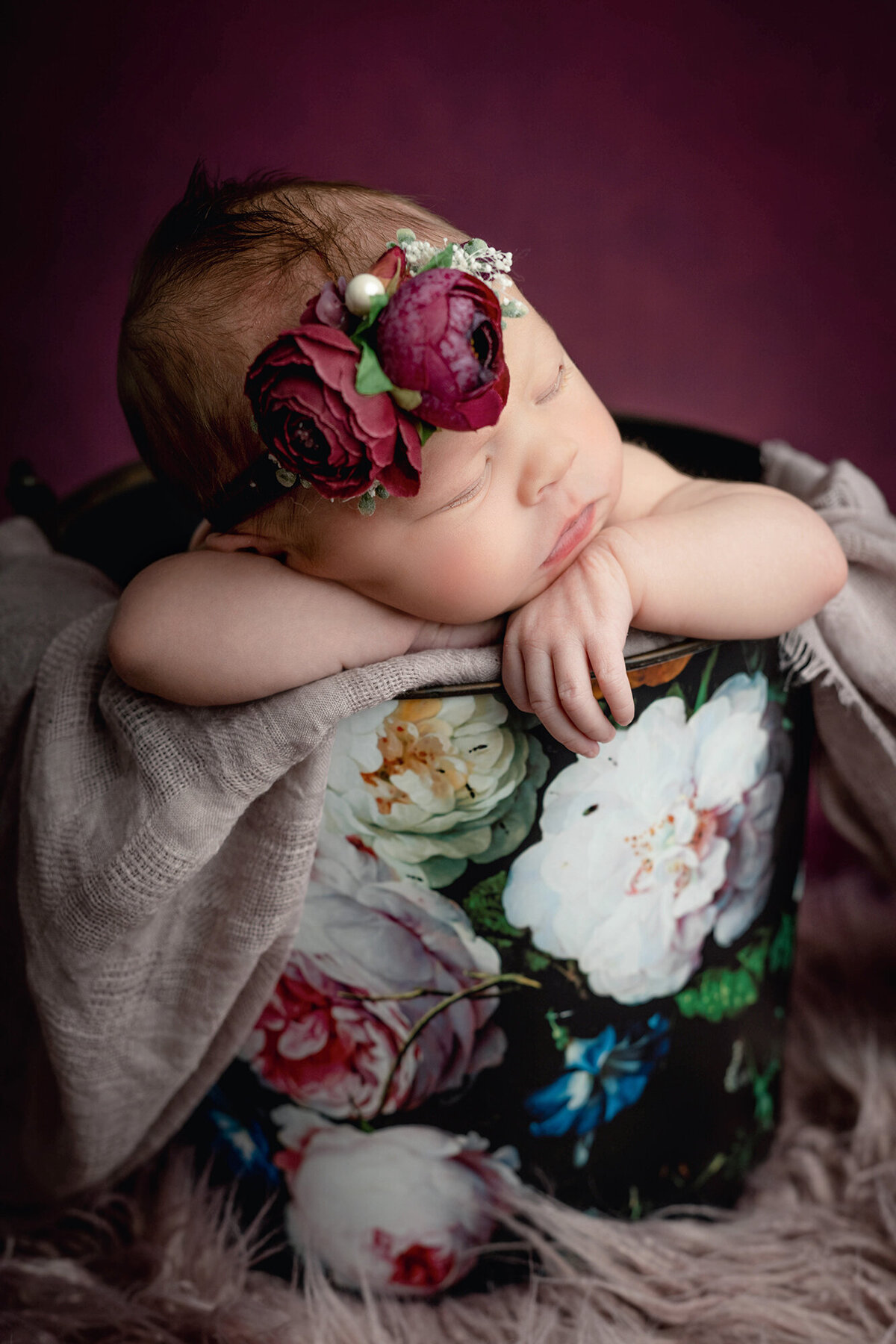 A newborn girl sleeps on the rim of a floral print bucket with a matching floral headband