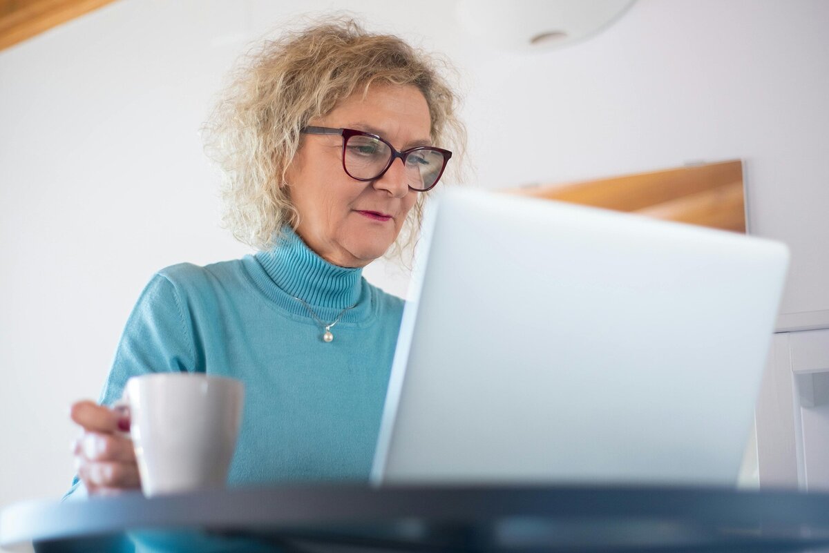 Middle Aged White Woman in Blue Sweater & Glasses Looks at a Laptop With a Cup Of Coffee Next To Her