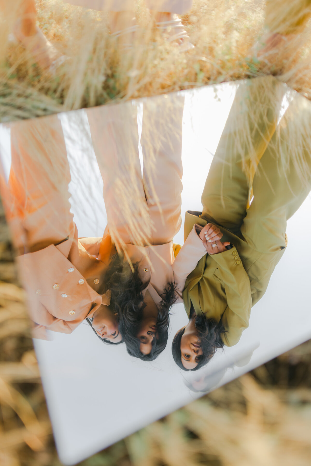 A surreal reflection of three women in pastel and earth-toned suits captured through a mirror placed in dry grass, creating an artistic and conceptual editorial feel. Greenville South Carolina portrait photographer – Dani Lacey Photographs.