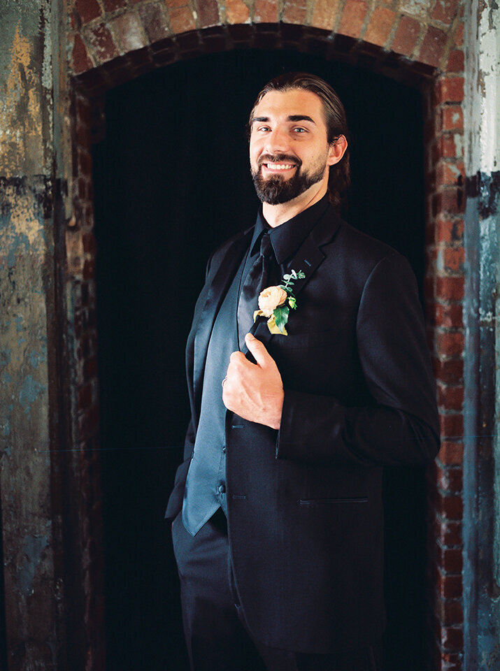 A groom wearing a black tuxedo smiles holding his jacket in front of a doorway