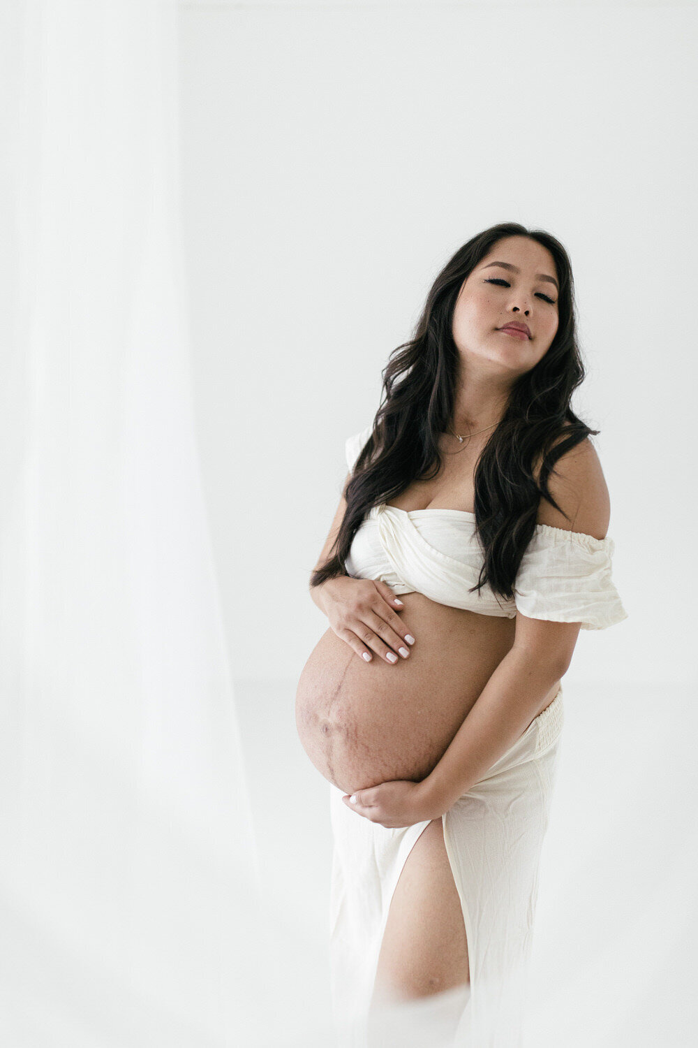 Woman stands in white room, wearing white and holds her pregnant bump whilst looking over her shoulder