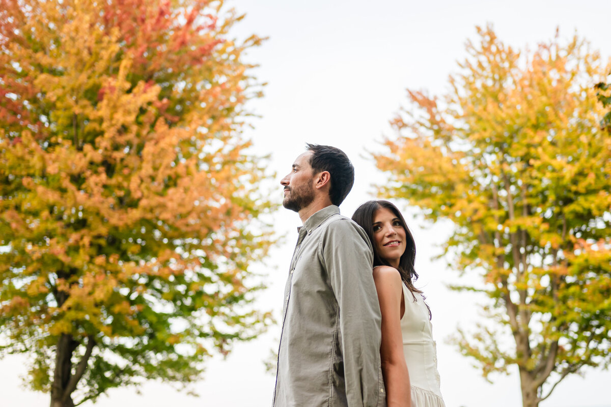 A couple stand back to back in front of fall tree