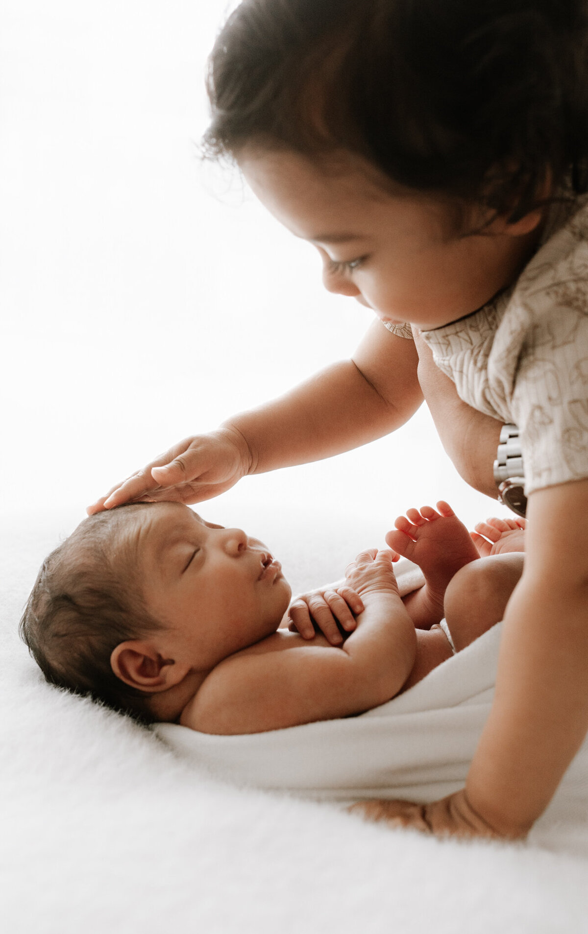 A tender moment between siblings during a newborn photography session. A toddler gently places their hand on their sleeping newborn sibling’s head, showing curiosity and affection. The newborn is wrapped in a white blanket, peacefully resting on a soft surface. Captured in natural, diffused light at a Sydney photography studio, this image beautifully documents the beginning of a sibling bond.