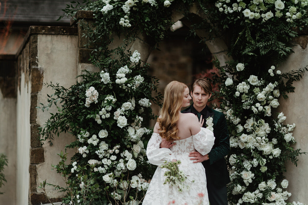 A bride and groom embrace during their bridal portraits in a summer arch of white roses and florals