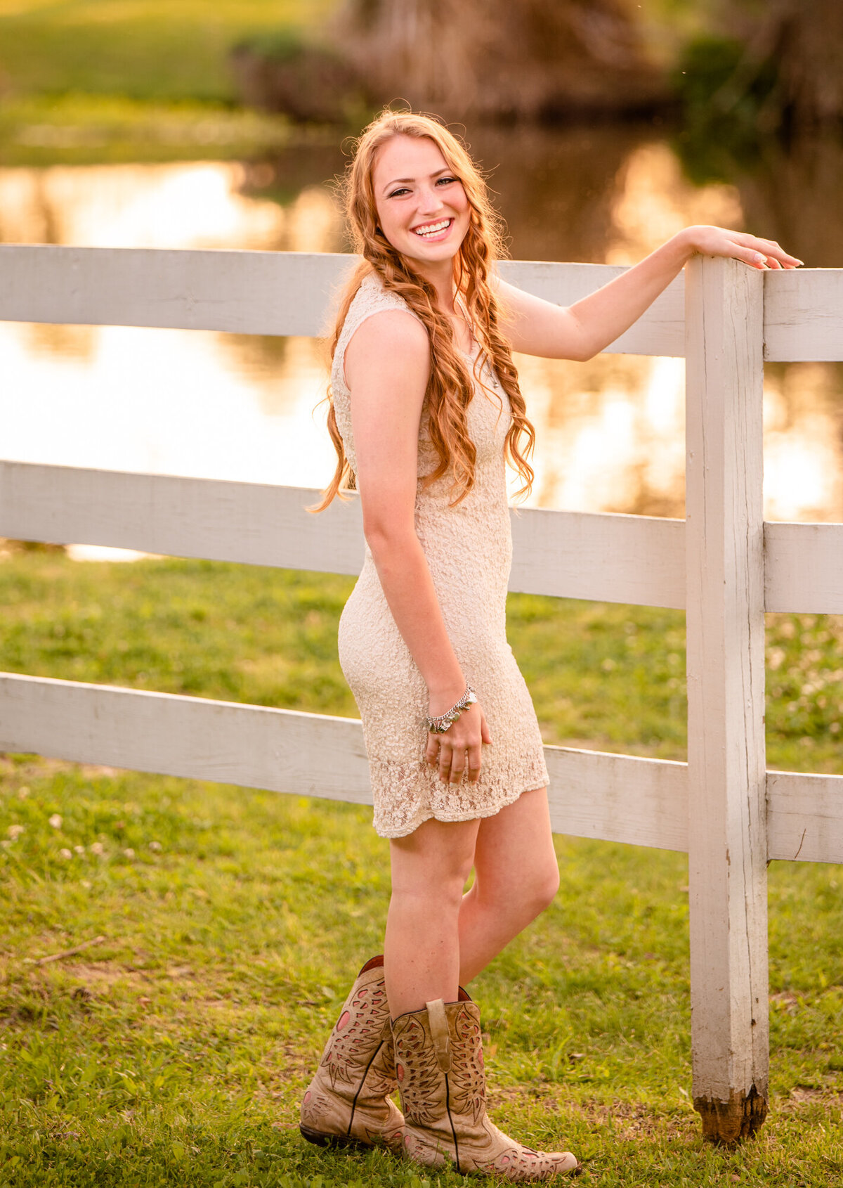 A senior wearing a white dress and cowboy boots stands next to a white fence and smiles at the camera.